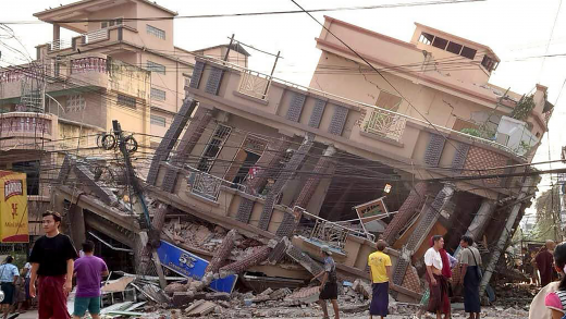 Een ingestort gebouw in Myanmar. De hele fundering is weg, mensen kijken er verloren naar. 