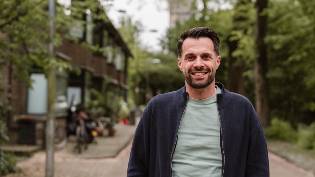 Jurjen ten Brinke staat in een straat van een woonwijk in Amsterdam Noord. Hij kijkt lachend de camera in.