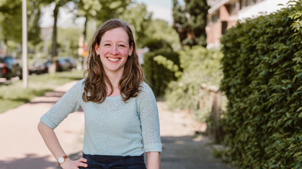 Simone staat in een woonwijk en kijkt lachend de camera in. Ze draagt een mintgroene top. Op de achtergrond zie je huizen en een fietspad.