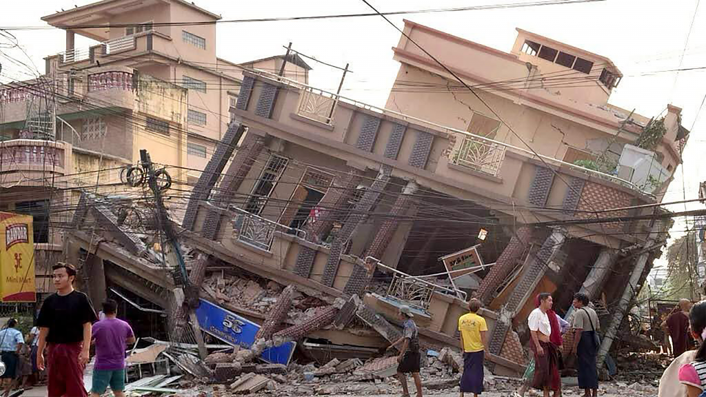 Een ingestort gebouw in Myanmar. De hele fundering is weg, mensen kijken er verloren naar. 