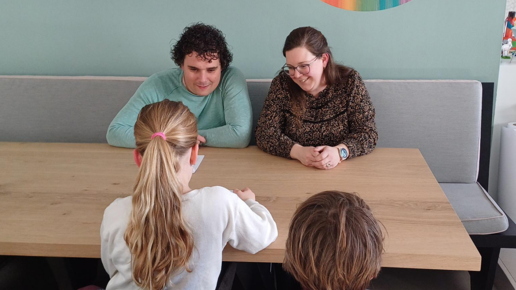 Twee ouders zitten met hun twee kinderen lachend aan tafel terwijl een kind voorleest uit de veertigdagenkalender.