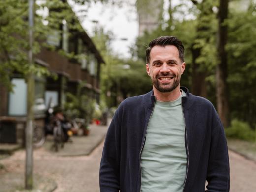 Jurjen ten Brinke staat in een straat van een woonwijk in Amsterdam Noord. Hij kijkt lachend de camera in.