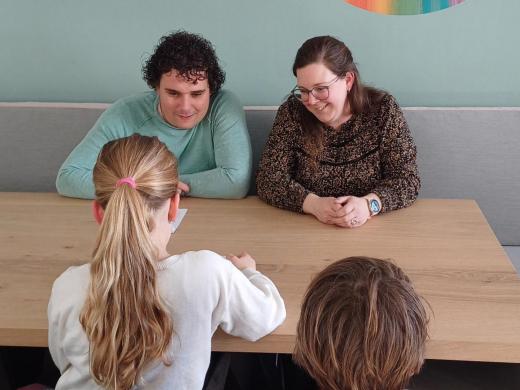 Twee ouders zitten met hun twee kinderen lachend aan tafel terwijl een kind voorleest uit de veertigdagenkalender.