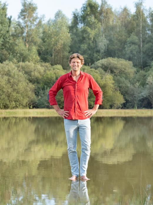 Alfred Slomp staat met blote voeten op een waterplas. Hij draagt een felrood overhemd, heeft zijn handen in zijn zij en kijkt de camera in.